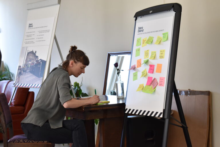 An image of a member of the public writing their feedback at the public consultation. Two whiteboards show images of the Fabric District and post-its written by members of the public.